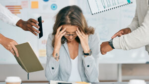 A woman business owner holds her head in her hands. Around her, someone is pointing at a watch, someone is handing her a phone, and someone is pointing to a journal.