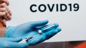 A medical professional draws a vaccine from a vial. Behind is a sign reading, "COVID19."