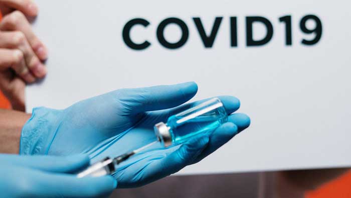 A medical professional draws a vaccine from a vial. Behind is a sign reading, "COVID19."