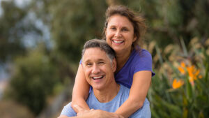 A senior woman hugs her husband from behind. They both smile.