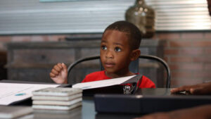 A young boy sits at a desk with paperwork in front of him. He looks to someone off screen for guidance.