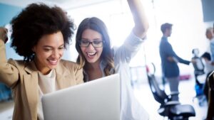 Two women celebrate something they see on their laptop screen.
