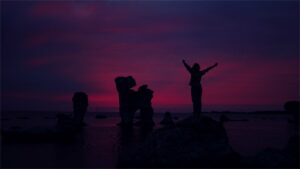 A group of friends sits on a dock at night. One stands with their arms open wide joyfully.