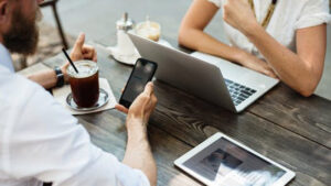 Two people discuss insurance while at a coffee shop. One is on their phone, and the other is on a laptop.