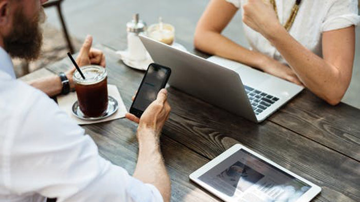Two people discuss insurance while at a coffee shop. One is on their phone, and the other is on a laptop.