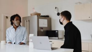 Two people wearing masks discuss work while social distancing. There is a laptop open.