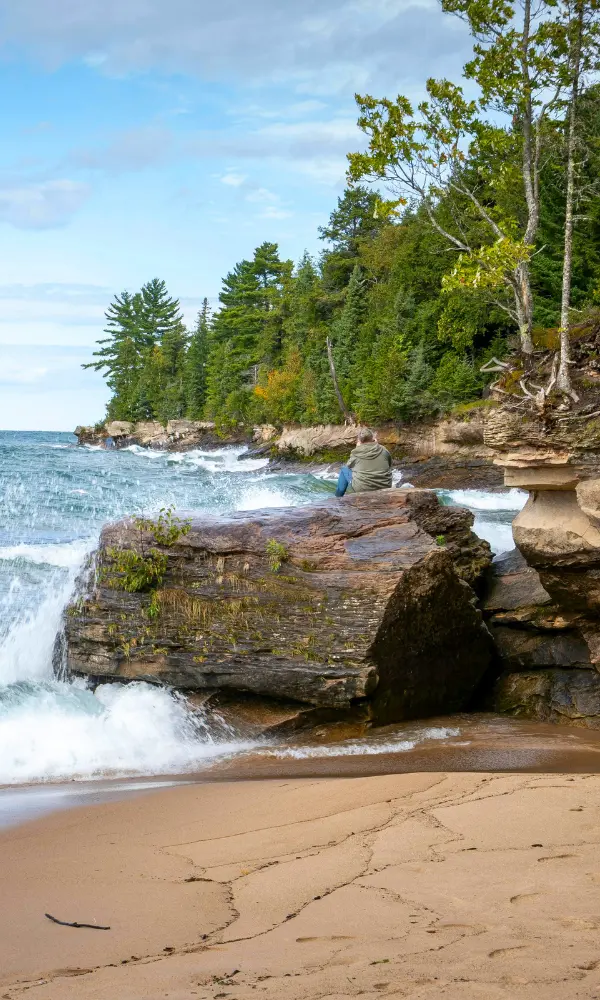 A person sits on a large rock on a beach in Michigan.
