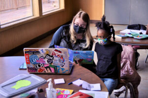 A volunteer with the Charles Stewart Mott Foundation reads to a girl. They both wear masks. The foundation supports the Flint community by supporting education and through many other efforts. (Image via the Charles Stewart Mott Foundation website)